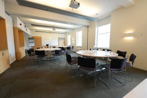 A modern conference room in a flexible event space in London features round tables and purple chairs arranged in a semi-circle. A projection screen displays a presentation, while large windows flood the room with natural light. Water bottles and glasses are placed on tables, complementing the breakout catering area.