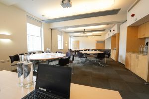 A modern, sustainable conference events room in London features round tables and chairs, bottles of water, and a laptop on a table in the foreground. The spacious room boasts large windows for natural light and a ceiling-mounted projector, ideal for medium-sized events.