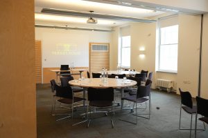The sustainable events venue in London features a conference room with round tables and chairs in a meeting setup. Water bottles and glasses adorn the tables, while a projector displays Friends House on the screen. Large windows and neutral-colored walls complete this flexible event space near London Euston & Kings Cross.