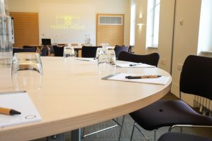 A round conference table with notepads, pens, and water glasses is set up in a meeting room. The screen in the background highlights Friends House in London Euston & Kings Cross, showcasing a flexible meetings events suite surrounded by several empty chairs.